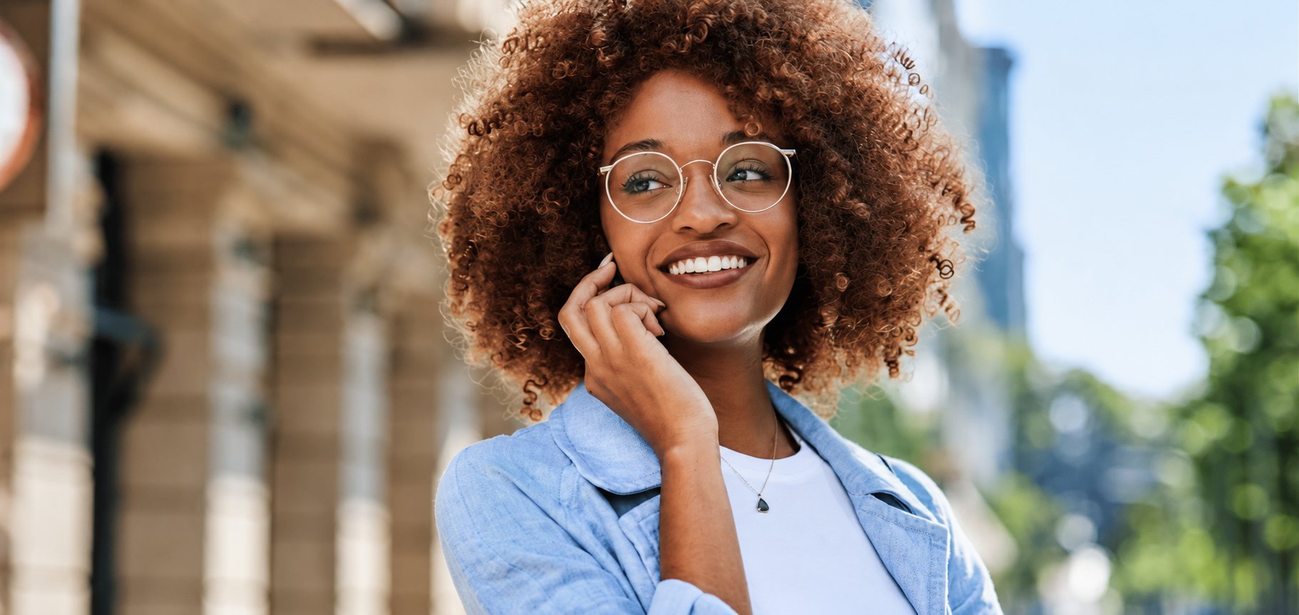 Femme souriante aux cheveux bouclés et lunettes rondes, au téléphone en extérieur